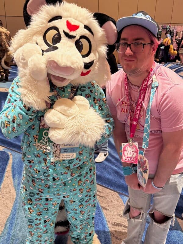A person in a colorful, fluffy tiger costume with prominent eyes poses beside another person wearing glasses, a blue cap, and a pink shirt. The costume is light blue with various cute patterns, including stars and rainbows. The person in the costume is making a playful gesture with their paw to their cheek. The person beside them has a pink lany