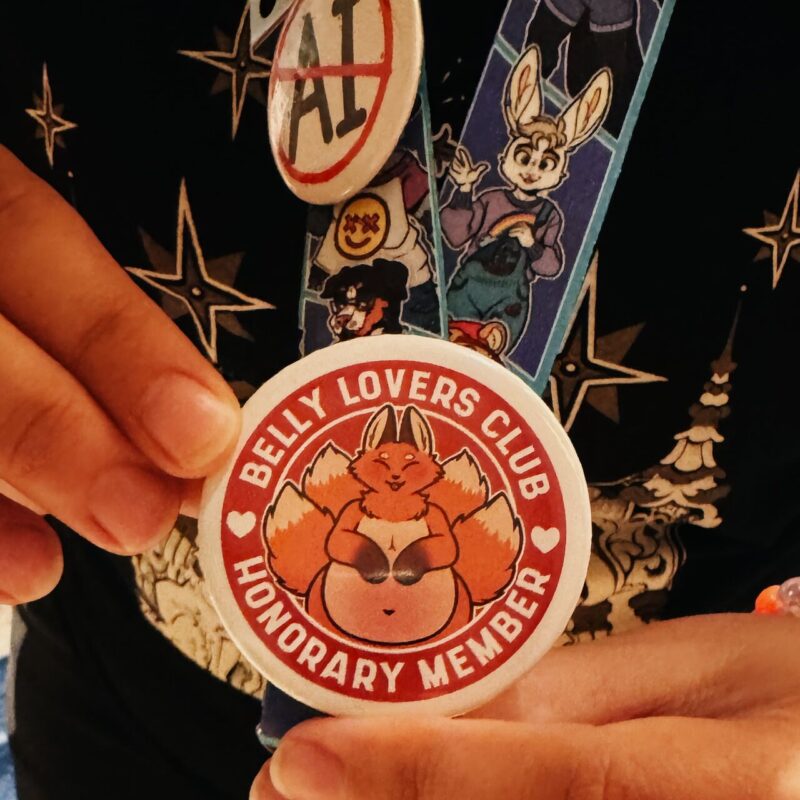 A person holds a circular badge with the words BELLY LOVERS CLUB HONORARY MEMBER in white against a red background. The badge features an illustration of a smiling, chubby fox with multiple tails. The person is also wearing a lanyard with colorful cartoon characters and another button displaying No AI crossed out. The clothing has