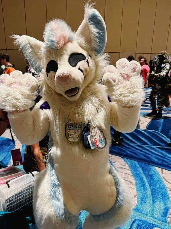 A person wearing a fluffy, white and pastel blue anthropomorphic animal costume stands indoors on a patterned carpet. The costume features large ears, a tuft of pink fur on top of the head, black markings around the eyes, and pink paw pads. Badges are hanging around the neck. Behind, other people in costumes are visible, along with a large