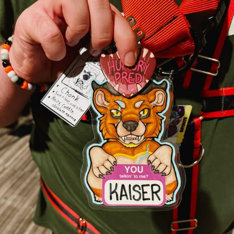 A close-up image of a hand holding a colorful badge that features an illustrated anthropomorphic lion with a fierce expression and large fangs. The badge includes text that reads YOU talkin' to me? and KAISER. Above it, there's a heart-shaped pin with the words HUNGRY PRED. The person is wearing