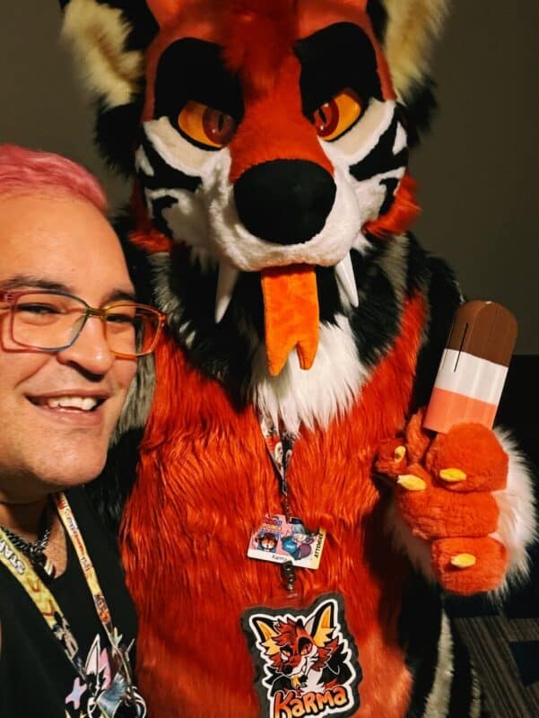 A person with pink hair and glasses is taking a selfie with someone in a furry costume. The costume is red, black, and white, resembling a tiger or wolf with large orange eyes and an orange tongue sticking out. The character holds a popsicle prop with brown and white layers. Both the person and the costumed character wear colorful lanyards