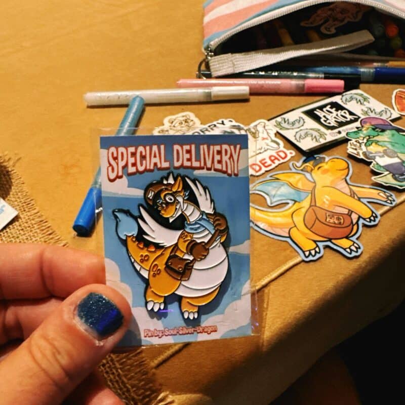 A person holds a colorful pin with a cartoon dragon dressed as a mail carrier, featuring a bag and goggles, against a sky-blue background. The words Special Delivery are written above the dragon. In the background, a table is scattered with various art supplies, including pens, markers, and additional stickers of cartoon characters. A striped pencil case is