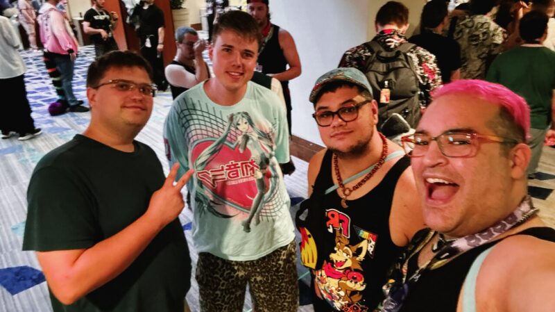 A group of four people happily pose for a selfie indoors at a lively event. The person on the right has pink hair and is smiling broadly. The person next to him is wearing a cap, glasses, and a tank top with colorful designs. The person in the center wears a t-shirt featuring an anime character print. The person on the left,