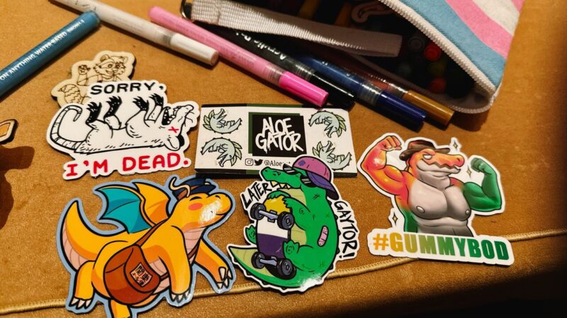 A collection of colorful stickers is spread on a surface next to a partially open pencil case with markers visible. Stickers include a cartoon possum playing dead with the text SORRY I'M DEAD, a cartoon dragon carrying a satchel, and various playful alligator-themed designs, including one flexing muscles with #GUMMYBOD text.