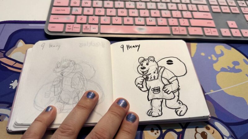 A sketchbook open to two pages is depicted. The left page shows a rough pencil sketch, and the right page features a detailed ink drawing of an anthropomorphic bear carrying a bag labeled 9 Heavy. The bear is dressed in overalls with a coffee cup symbol. The sketchbook rests on a colorful desk mat, and a person's hand with shiny nail polish