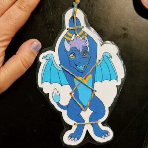 A hand with lavender-painted nails is next to a laminated paper cutout of a cartoon dragon on a dark surface. The dragon is blue with purple hair and yellow eyes, featuring wings and a playful expression. It is decorated with rainbow-colored string threaded through holes in the cutout, creating a pattern across the dragon’s body. The dragon appears to be