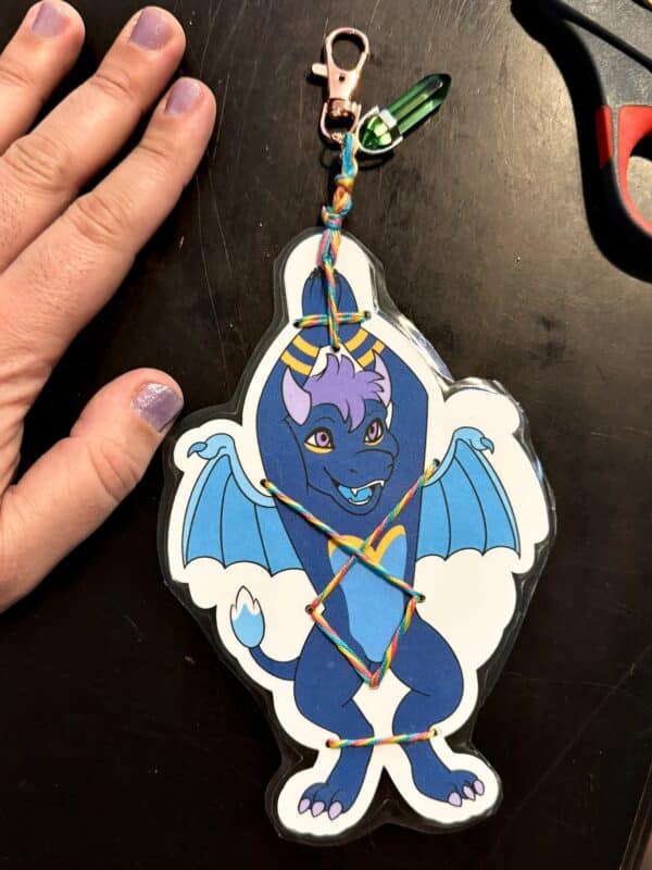 A hand with nail polish rests on a table next to a colorful dragon keychain. The dragon is blue with purple accents and wings, and it appears cheerful. A multi-colored string is artfully laced across its body. The keychain has a metallic clasp and a green crystal charm attached. Scissors with red and black handles can be partly seen
