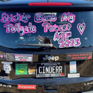 The image shows the rear of a black Jeep vehicle covered with various stickers and colorful writing. The message in pink and white paint on the window reads, Sticker Bomb My Tailgate Pwease AQF 2025 with a heart symbol. Stickers include phrases like Hot single werewolves in your area?, My Other Ride Is A