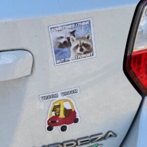 A car's rear end with two stickers. The upper sticker features two raccoons with the text Sometimes I think but then I forget. Below, a sticker shows a dog riding a toy car, captioned Vroom Vroom. The word Impreza is partially visible below the stickers. The car's brake light is on