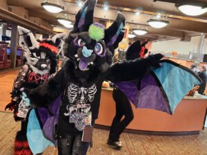 Two people wearing furry costumes pose in an indoor setting. The costume in the foreground features a black and purple bat-like design with green and blue accents, and visible wings. A skeleton t-shirt is visible, along with various badges and tags. Behind, another individual wears a black, white, and red wolf-like costume. The environment suggests a public space