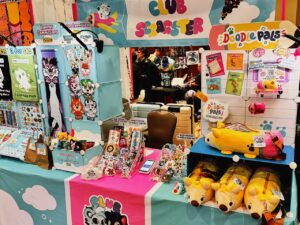 A colorful vendor booth at a convention features cartoon-themed merchandise. Various items are displayed, including plush toys shaped like animals, keychains, enamel pins, and art prints. Two banners, Club Starster and Doodle Pals, hang overhead. The table has bright tablecloths with cloud patterns and the display is vibrant with character illustrations