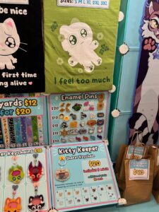 The image shows a colorful display at a merchandise booth. A green T-shirt with a cute, wide-eyed character and the words I feel too much is prominently featured. Below, various enamel pins are priced between $10 and $20. Keychains, labeled Kitty Keeper Shaker Keychain, are priced at $20, with additional