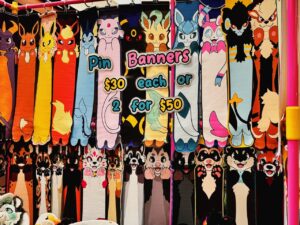 The image displays a colorful arrangement of cartoon-style pin banners featuring various long, vertically extended characters. Each character is distinct, depicting different animals or creatures with vibrant colors. The banners are displayed in two rows, and a sign reads, Pin Banners $30 each or 2 for $50 in playful font. The scene is lively, with