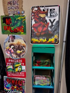 The image shows a display of colorful metal signs and trays. The signs feature various designs, including a red dragon, a green creature with No Smoking, and a dinosaur on a Private Property warning. There's also a circular fantasy-themed sign and a vibrant Beware Dog sign. A small shelving unit holds trays with similar designs, including
