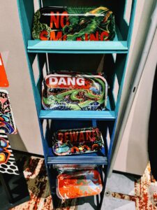 A teal shelf displays four colorful trays, each with vivid artwork. The top tray features a green dragon and the partial text NO SMOKING. Below, a tray with a lizard suggests the word DANG. The third tray showcases a cartoonish blue dog with the text BEWARE of DOG. The bottom tray is red with abstract
