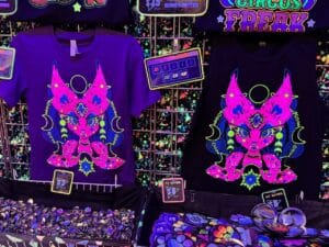 A vibrant display features two shirts with psychedelic, neon designs of a mystical, feline-like creature adorned with celestial symbols. The left shirt is purple, and the right is black, both with striking pink, green, and blue details. Below, a variety of colorful buttons are spread out, each priced at $2. The background is a grid with spl