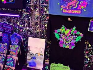 A vibrant display of colorful merchandise featuring neon-themed designs. A black T-shirt showcases a cartoon bat with bright green wings and pink accents, labeled “Bat Crazy.” Above it, a blue shirt displays a skateboarding graphic with the words “Big Cat Summer.” To the left, various patches are organized on racks, each with distinct bright illustrations. A small