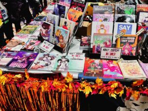 A table displaying a wide array of comics and graphic novels with colorful covers, including illustrations of animals and humans. Many are marked as adult content, 18+ only, with prominent signs requesting ID verification. The table is decorated with autumn-themed ornaments, such as orange and yellow leaves, creating a festive look. Various titles and artwork are visible, with
