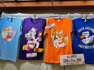 Four colorful T-shirts are hanging side by side. The first shirt is blue with a playful character holding a cup. Next is a purple shirt featuring a cheerful white cat with a volleyball. The third shirt is orange, displaying a white animal character. The last shirt is blue with a duck and text that reads, DUCK AROUND AND FIND OUT
