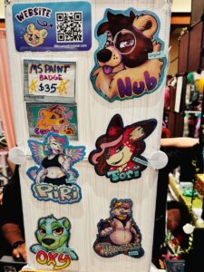 This image shows a display board with various cartoon-style animal character badges at a convention booth. The badges feature colorful illustrations of anthropomorphic animals with names like Nub, Piri, Tori, Oxy, and Tiramisu. Each character has a unique design and vibrant colors. The top left corner has a website QR