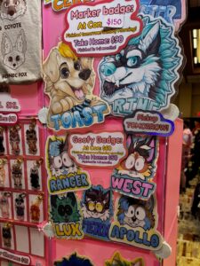A colorful display at a convention features cartoon animal character badges and keychains. The top section has Marker badges showing a playful dog named Toast and a wolf named River. Prices are listed: $150 at the convention, $90 to take home with approximately 1-2 months for completion. The middle section shows Goofy