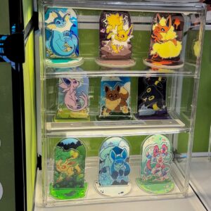 A display case features colorful, stylized acrylic standees of various Pokémon characters, arranged in three rows. The top row shows a blue aquatic creature, an electric yellow one, and a fiery orange one. The middle row includes a psychic purple character, a brown fluffy one, and a dark, mysterious black one with rings. The bottom row contains a