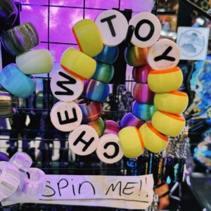 Colorful, segmented toy bracelet hangs on a display, consisting of multicolored beads and white beads with black letters spelling NEW TOY. A handwritten sign below reads SPIN ME! The background is lit with purple and reflects various other toys and objects in a shop-like setting.