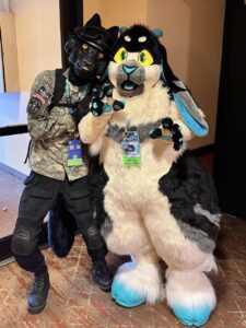 Two people wearing costumes pose together for a photo. One is dressed in a black animal suit with yellow eyes and military-style clothing, including a helmet and patches. The other wears a large, fluffy costume resembling an animal with black, white, and blue fur, featuring bright yellow eyes, blue accents, and long ears. Both have badges around their neck