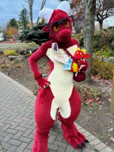 A person in a red and white dragon fursuit poses confidently in an outdoor park setting. The costume includes large horns, black glasses, and expressive orange eyes. They wear a crossbody bag embellished with character badges and hold a small plush toy. The ground is paved with a brick pathway, surrounded by bare trees and sparse greenery, suggesting a fall