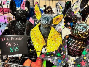 Colorful stuffed bats are displayed in a metal grid basket. Each bat has unique fabric patterns, including starry black, bright yellow with designs, and multicolored with stars. A chalkboard sign reads It's frickin' BATS: $45 in pink and green lettering. The playful assortment suggests a whimsical craft or toy stall setting