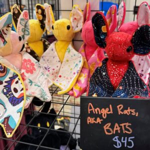 A collection of colorful plush bats displayed on a grid shelf. Each bat is designed with various patterns and colors, including pink, yellow, and red, with large ears and stitched features. Their wings showcase different whimsical patterns. A chalkboard sign beneath them reads Angel Rats, AKA BATS, $45 in orange and blue writing.