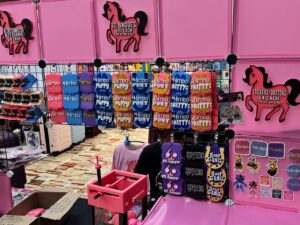 A pink retail display showcases various novelty items. The display features sections for retro computer keychains, 3D badges, and stickers/buttons with pricing. Keychains read phrases like Bimbo, Himbo, and Thembo in playful colors. There are also badges with phrases like Puppy, Pony, Kit