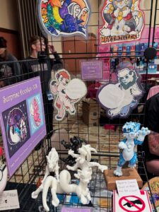 This image shows a display at a convention booth featuring various colorful, cartoon-style badges with anthropomorphic animal characters. The badges exhibit characters with speech bubbles and vibrant designs. Below, there are white and blue figurines of similar creatures, one set on a wooden base with a price tag indicating figurine commissions starting at $400. A sign offers Speech