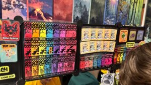 A display of colorful badges and cards at a booth. The left board features badges with various designs and text like CAUTION, WARNING, and WIDE LOAD in multiple colors. A Sold Out sign is visible. The right board shows badges with cartoon characters and phrases such as I'm baby! and I'm just a little
