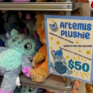 A display of colorful plushies is shown on metal shelves. The focus is on a light blue and gray plushie with large eyes and pink accents. Next to it is a sign with a drawing of a cartoon character labeled Artemis Plushie. The sign highlights features such as a magnetic mouth, beans in the feet and bottom, and compatibility