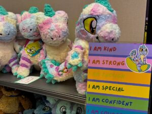 A group of colorful plush dinosaur toys is arranged on a shelf. The toys are pastel shades of pink, purple, blue, and green, with one prominently displaying a bandana. Beside them, a vibrant sign features affirmations in rainbow colors, saying I am kind, I am strong, I am enough, I am special
