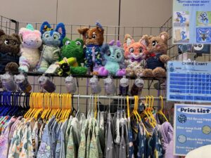 The image shows a display of colorful plush toys arranged on a metal shelf. The plushies include various cute animals, some wearing pajamas. Below them, racks hold various garments on hangers, featuring playful and colorful patterns. Beside the display, a chore chart with the name “Artemis” and a price list are visible. The price list