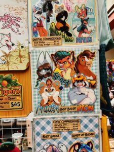 The image shows a display of colorful artwork featuring various anthropomorphic animal characters. There are several posters with characters in different poses, some having features like large ears and vibrant fur patterns. Text on the posters advertises Digital Commissions and Copic Commissions with prices listed for Bust Badge and Full Body Badge. The artwork