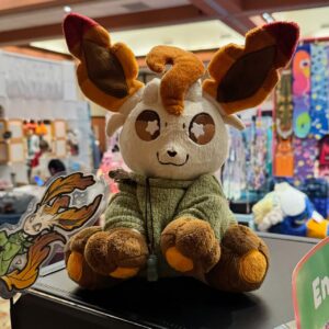 A cute plush toy featuring an animal-like character with large brown and orange ears, starry eyes, and a happy expression. It is wearing a green knitted sweater and has a small pendant around its neck. The plush is seated on a display, accompanied by an artistic illustration of the same character. In the background, there are colorful fabrics and products,
