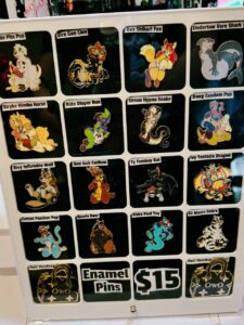A display board shows enamel pins featuring cartoon animals in various poses with playful expressions. There are four rows containing five pins each, labeled with names like Zira Goo Lion and Ava Shibari Fox. The styles are colorful and whimsical, showcasing different animals such as a lion, fox, shark, and snake. A lower section displays