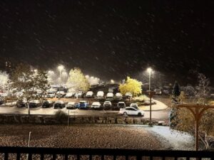 A nighttime scene showing a parking lot lightly dusted with snow. Snowflakes fall steadily against the dark sky, illuminated by several streetlights. A white car is driving along a road that curves through the parking area, which is lined with trees displaying some remaining autumn leaves. The ground and trees are lightly covered in snow, creating a wintry atmosphere.