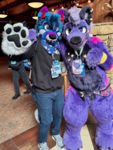 Two individuals in colorful furry costumes pose together indoors. The person on the left wears a blue wolf costume with a big paw raised, featuring vibrant blue and purple accents. They have a blue bandana around their neck. The person on the right wears a purple feline fur suit with colorful patterns and a vibrant fanny pack. Both have badges hanging from their
