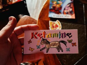 A person holds a sticker with the word Ketamine in colorful, stylized letters. Below the text, there is a cartoon illustration of a horse surrounded by colorful butterflies. The background is light purple. In the blurred background, a table with a partially visible book or magazine and another sticker featuring red and yellow designs can be seen.
