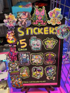 A black display board features colorful, illustrated stickers of various anthropomorphic characters. The board is labeled STICKERS $5 EACH in bright, neon colors. Stickers include characters with vivid, rainbow-hued designs and playful expressions. Some stickers have phrases like I'M FINE, LET'S GO CRAZY, and BAD DOG
