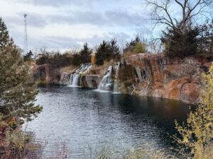 A scenic view of a tranquil waterfall cascading over rocky cliffs into a calm, reflective body of water. The landscape is surrounded by trees with sparse foliage, indicating late autumn or early winter. The foreground features some grasses and bushes, and a tall communication tower is visible in the distance on the left side. The sky is overcast, creating a serene and