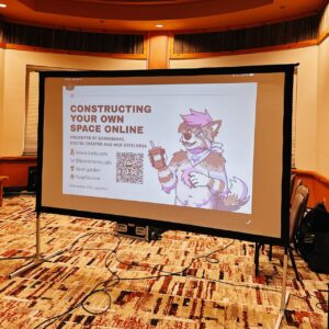A presentation screen displays a slide titled Constructing Your Own Space Online by a digital creator and web developer. Contact details and website links are shown alongside a stylized cartoon character of a wolf with pink hair, wearing glasses and holding a drink. The character sports a bandana and wristbands. A QR code is included. The room has beige