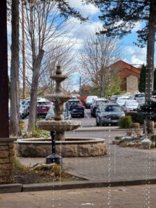 A picturesque outdoor scene features a stone fountain in the foreground with gentle water flow, surrounded by leafless trees. In the background, a parking lot is lined with various cars. To the right, there's a part of a red building with stone accents. Light snow dusts some grass and nearby surfaces, suggesting a chilly day. Strings of small lights add