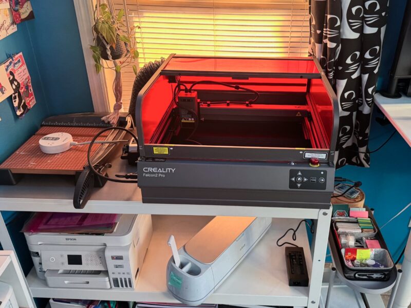 A Creality Falcon 2 Pro laser on top of a work bench in a home office, next to a paper cutter and above a printer and Cricut machine