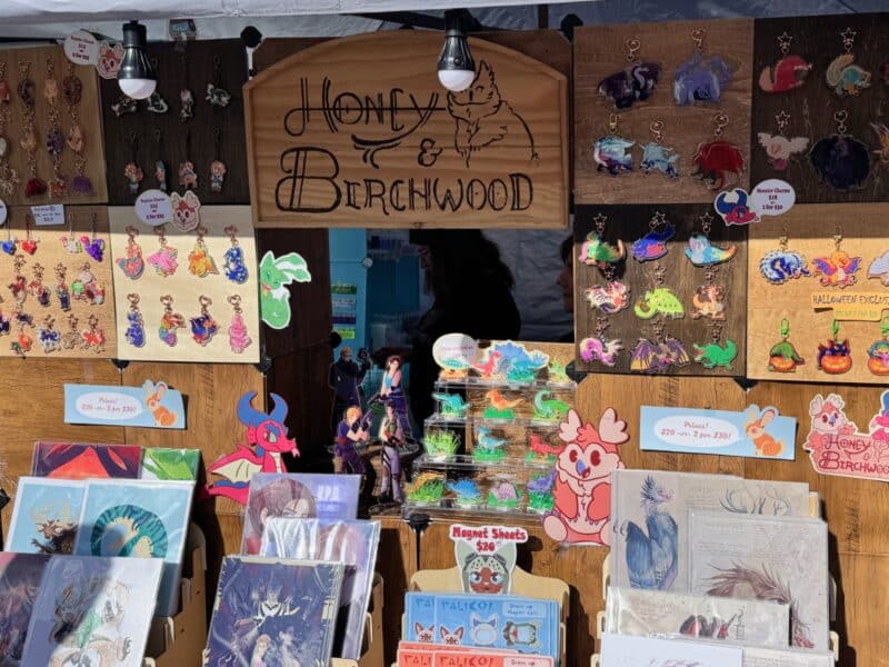 A booth at the Orlando Goblin Market for Honey and Birchwood, a shop with dnd and furry stickers and acrylic charms and prints on display