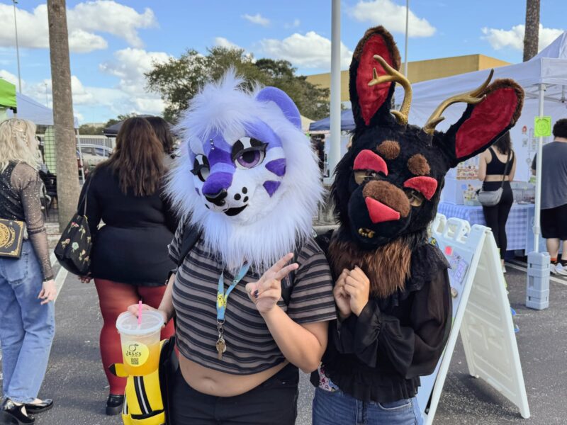 Two fursuiters at the Orlando Goblin Makret. One is a white cat with purple accents, and the other is a black jackalope with gold and red accents